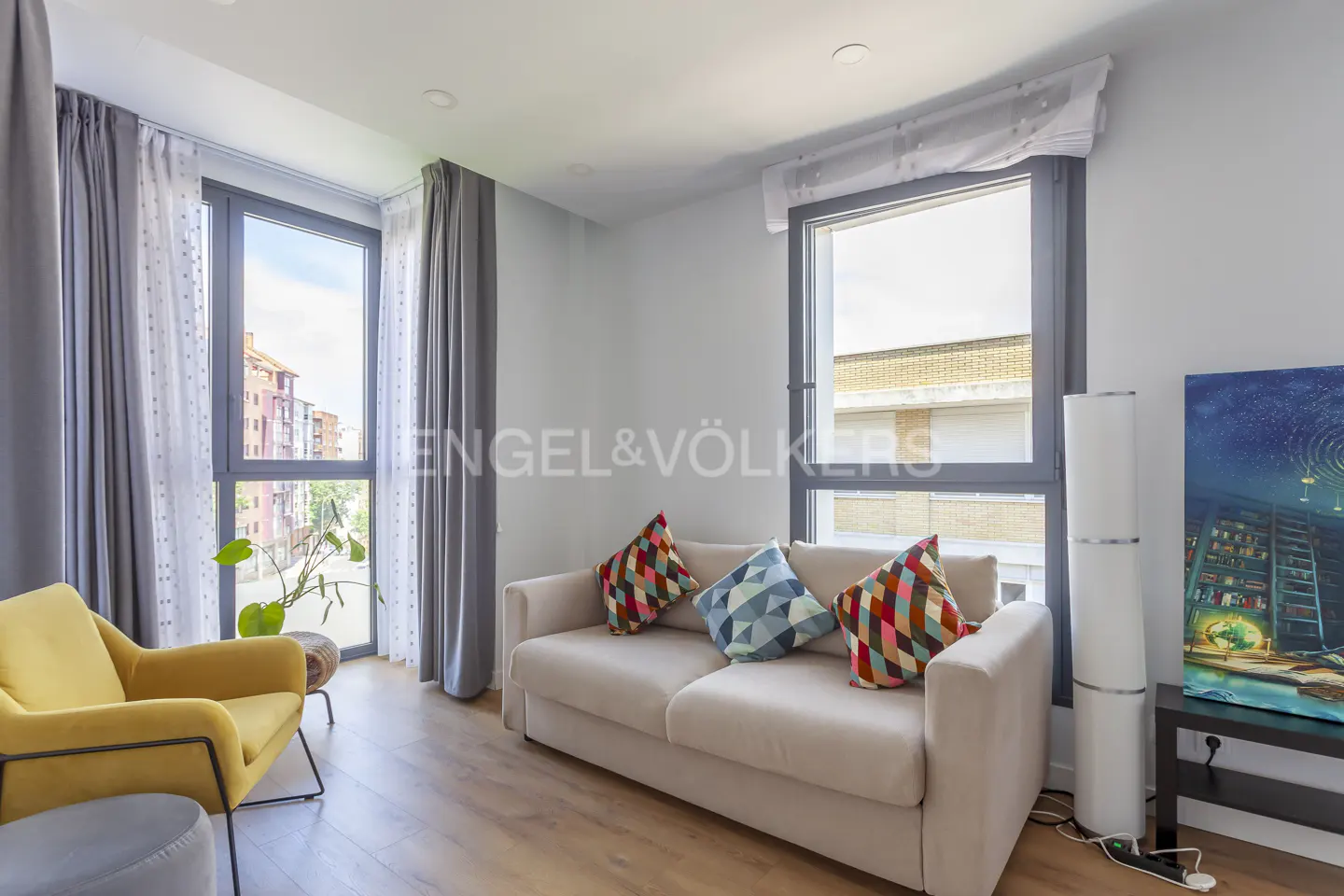 Modern living room with a beige sofa, colorful patterned pillows, a yellow armchair, and large windows with grey curtains.