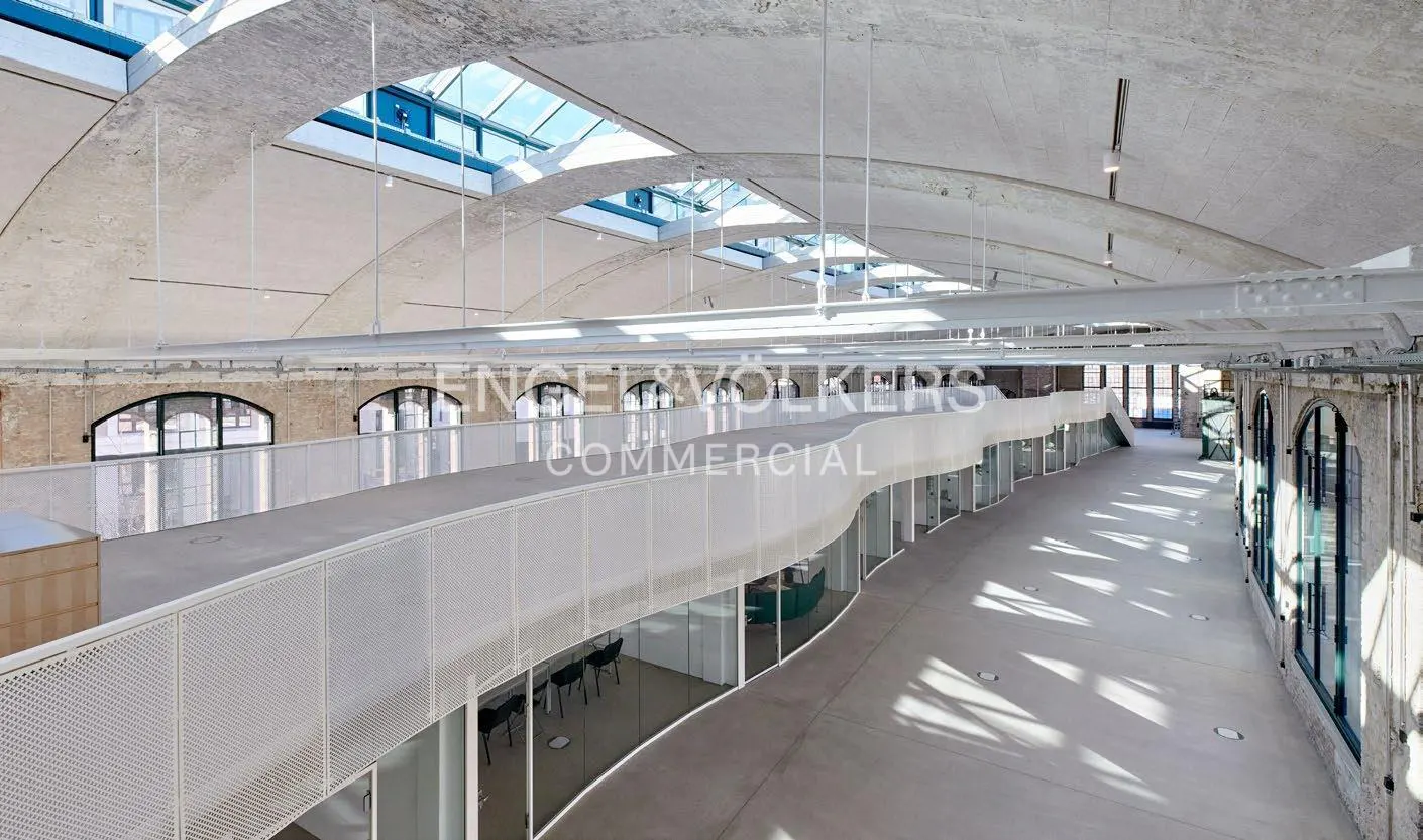 Interior view of a commercial space with a curved white balcony, glass-walled offices, and arched windows. Skylights illuminate the concrete ceiling.