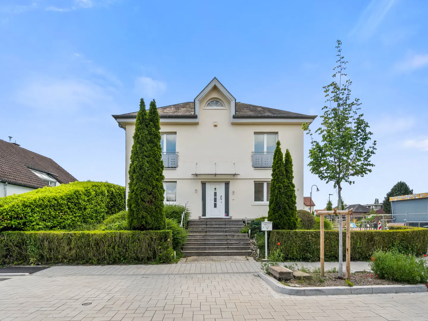 Two-story cream house with white trim, gray roof, and manicured green hedges under a blue sky.