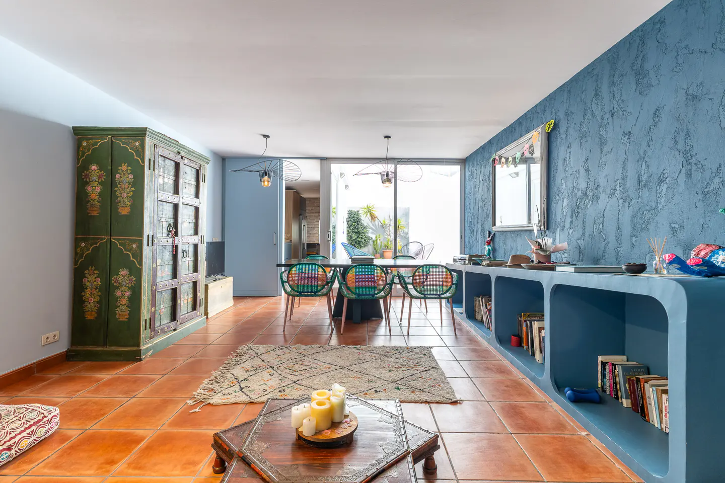 A bright living room with terracotta tile floors, a blue accent wall, and a green cabinet with floral designs. A dining table with colorful chairs sits near a large window.