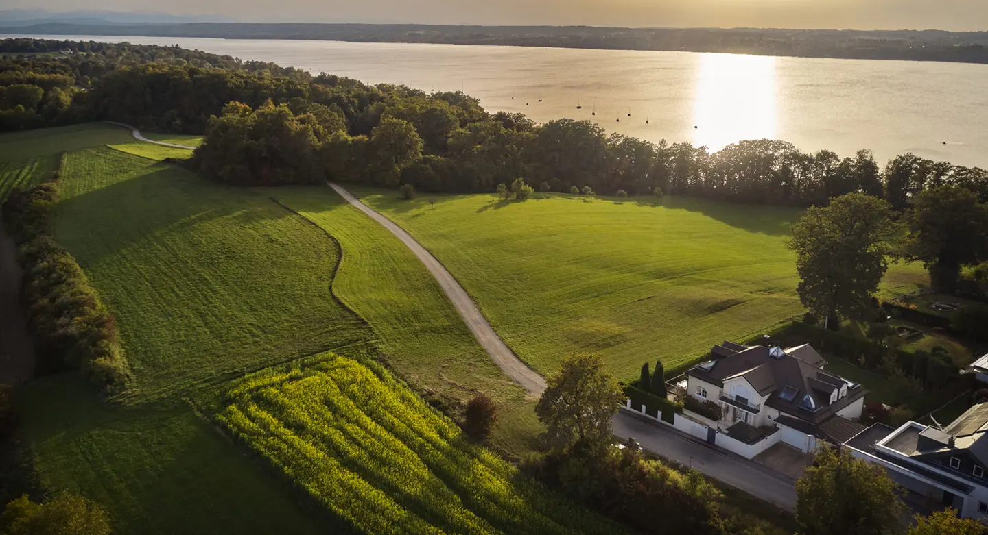 Aerial view of a modern white house near a lake, surrounded by green fields and trees under a bright sky.