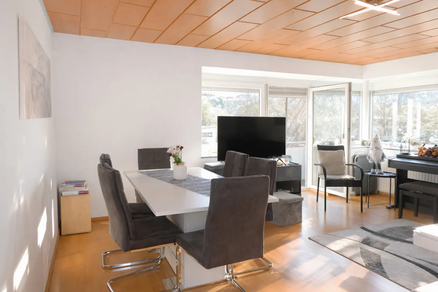Bright dining area with a white table, grey chairs, wood-paneled ceiling, and large windows overlooking the outdoors.