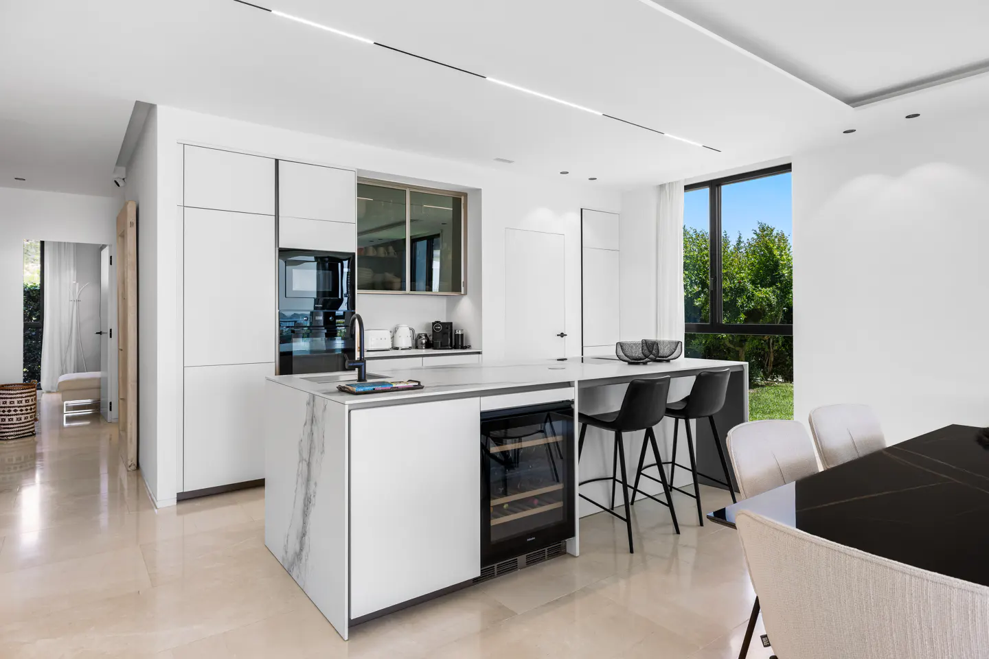 Modern white kitchen with a marble-accented island, black bar stools, built-in wine fridge, and a view of lush greenery.