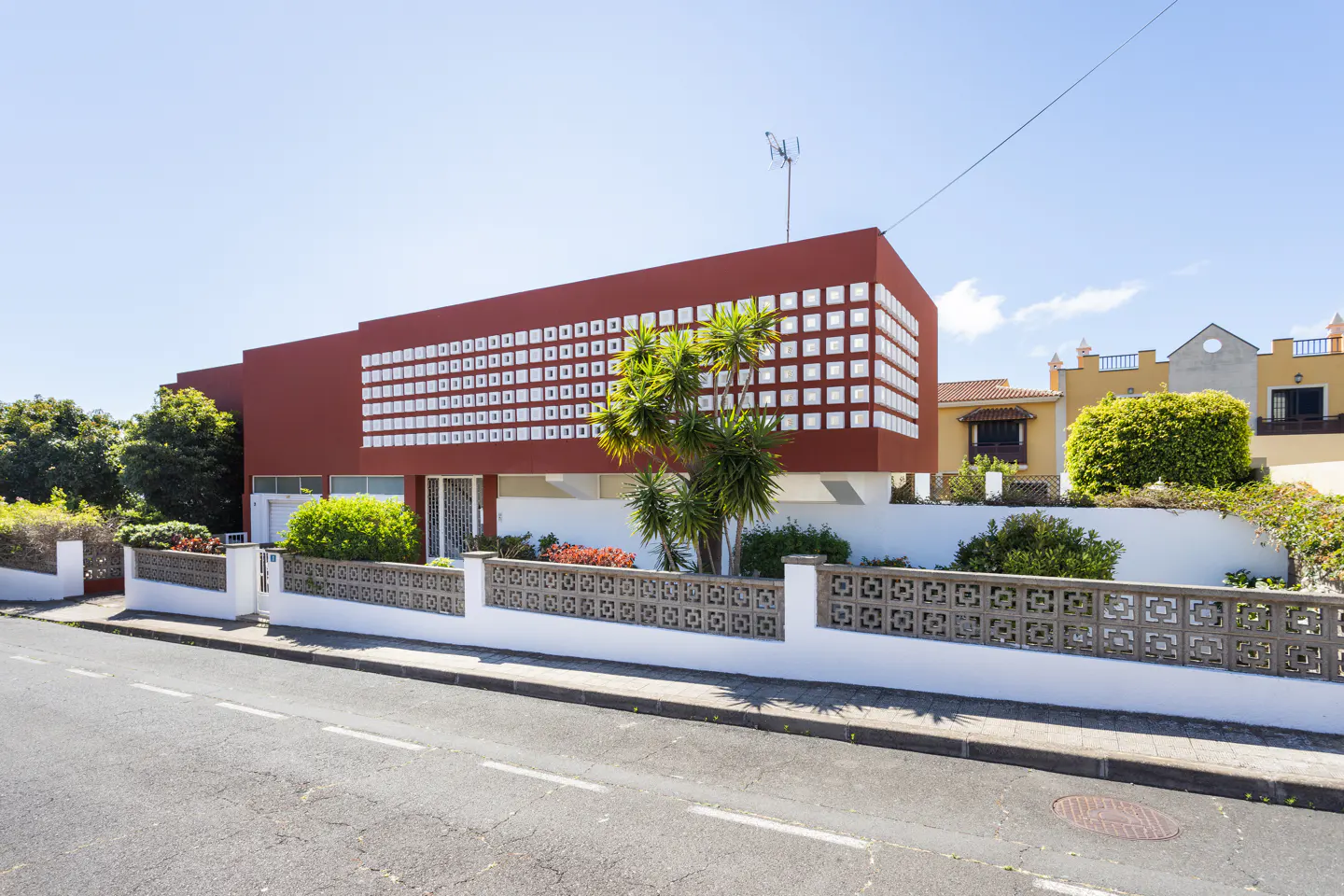 Modern red house with white square windows, a white and grey fence, and a palm tree in the front yard.
