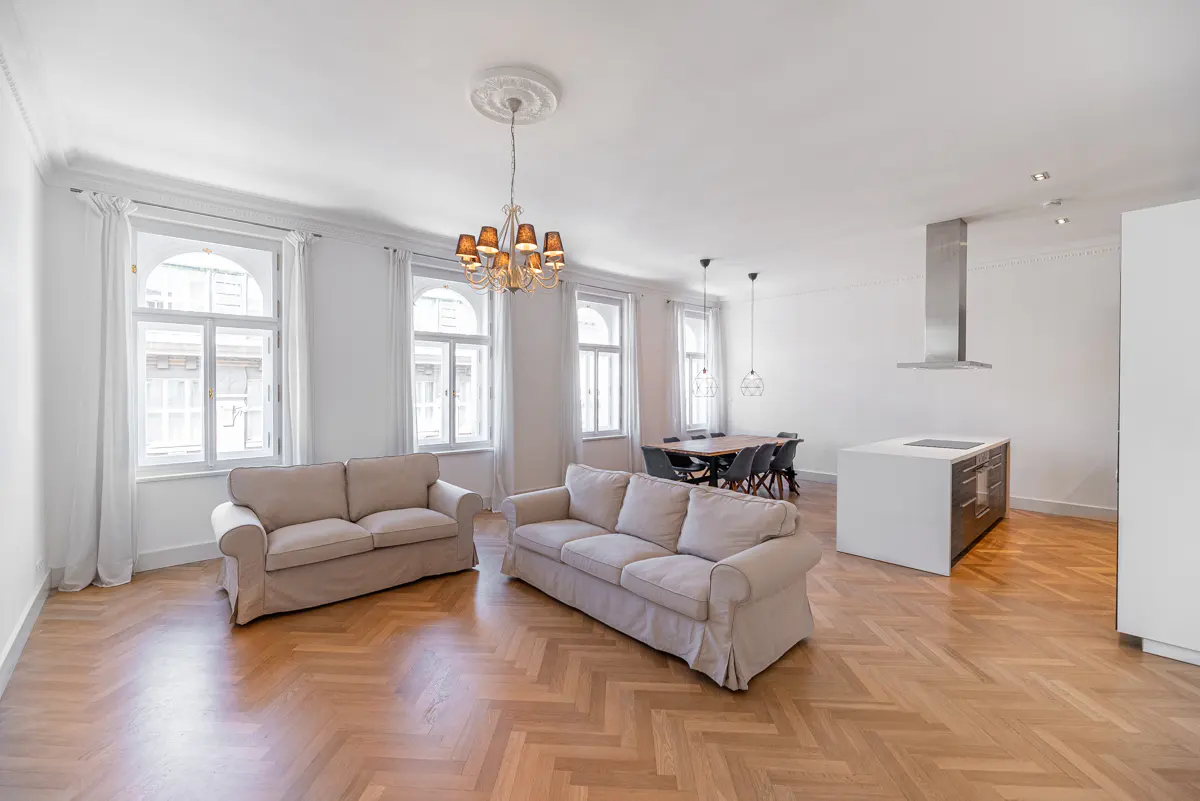 Bright, open-concept living space with two beige sofas, herringbone wood floors, and white walls. A dining table and kitchen island are visible in the background.