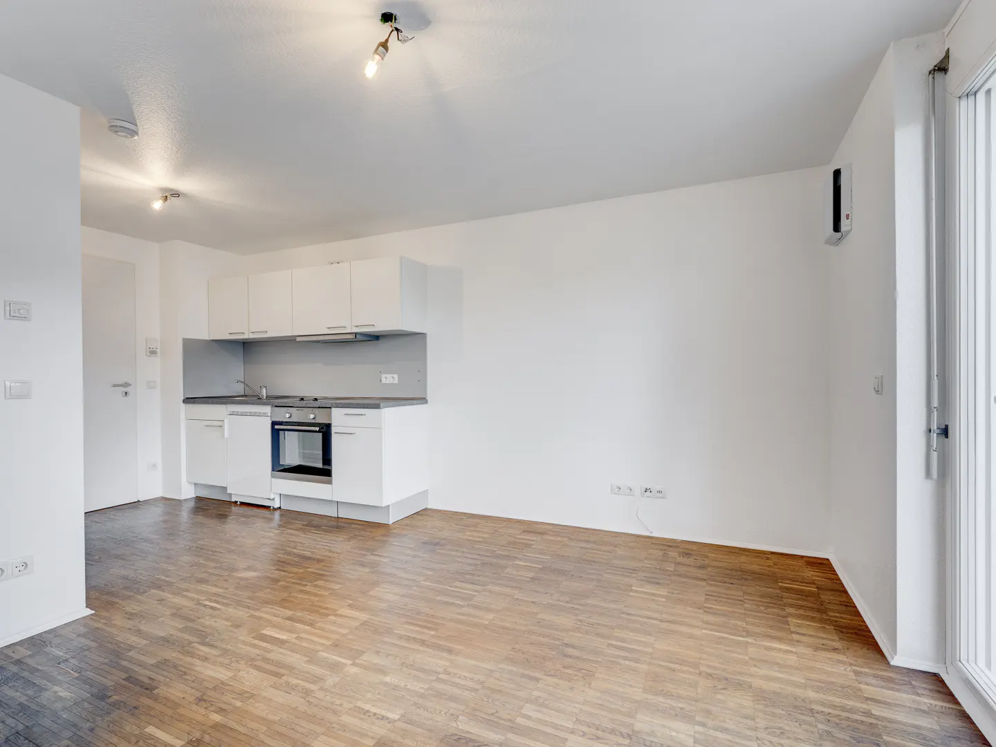 An empty apartment with white walls, a wood floor, and a white kitchen.