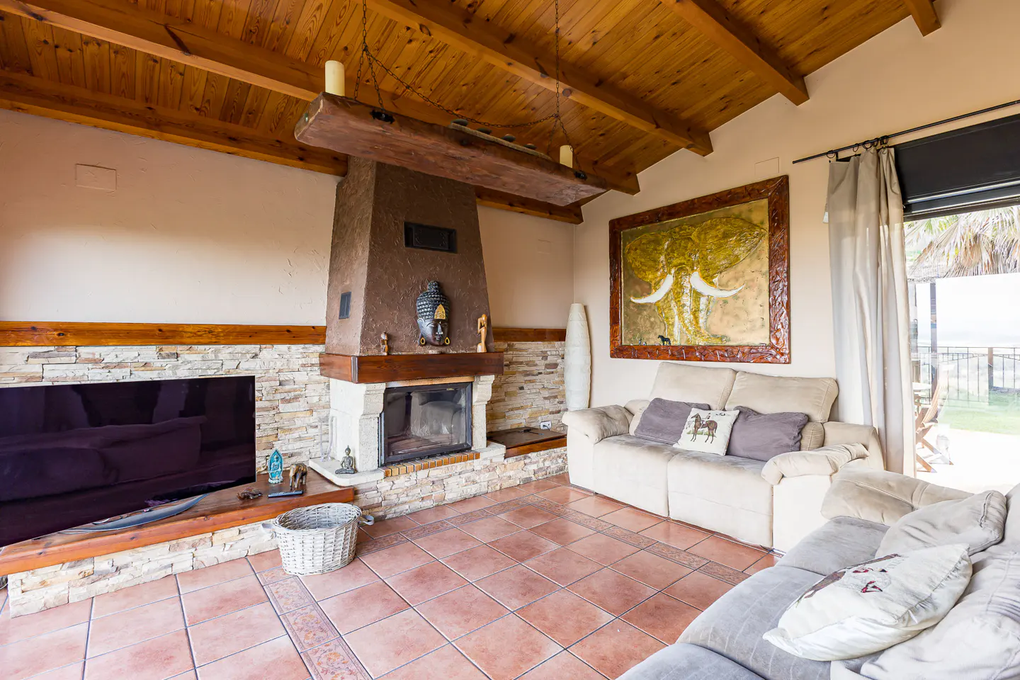 Living room with a stone fireplace, Buddha statue, wooden ceiling, beige sofa, and large elephant wall art.