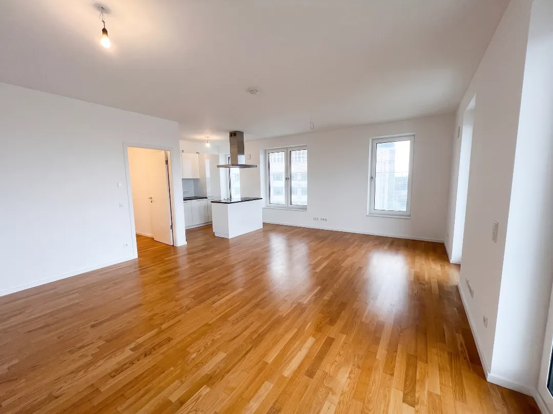 Bright, modern apartment with hardwood floors, white walls, and an open kitchen with a stainless steel range hood. Windows offer a city view.