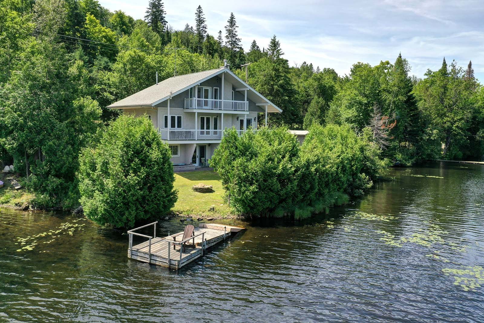 Two-story gray house with white balconies overlooking a lake with a wooden dock and chair. Green trees surround the house.
