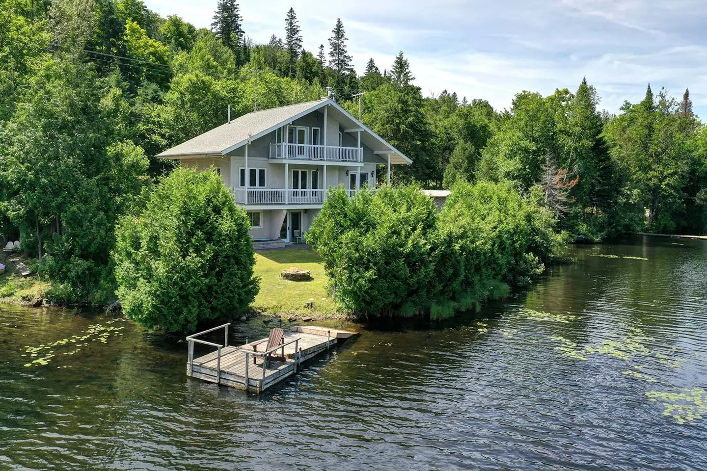 Two-story gray house with white balconies overlooking a lake with a wooden dock and chair. Green trees surround the house.