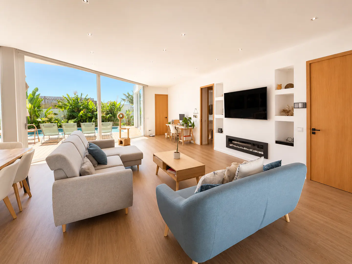 Bright living room with light wood floors, a blue sofa, a grey sectional, a fireplace, and a view of a pool and garden.