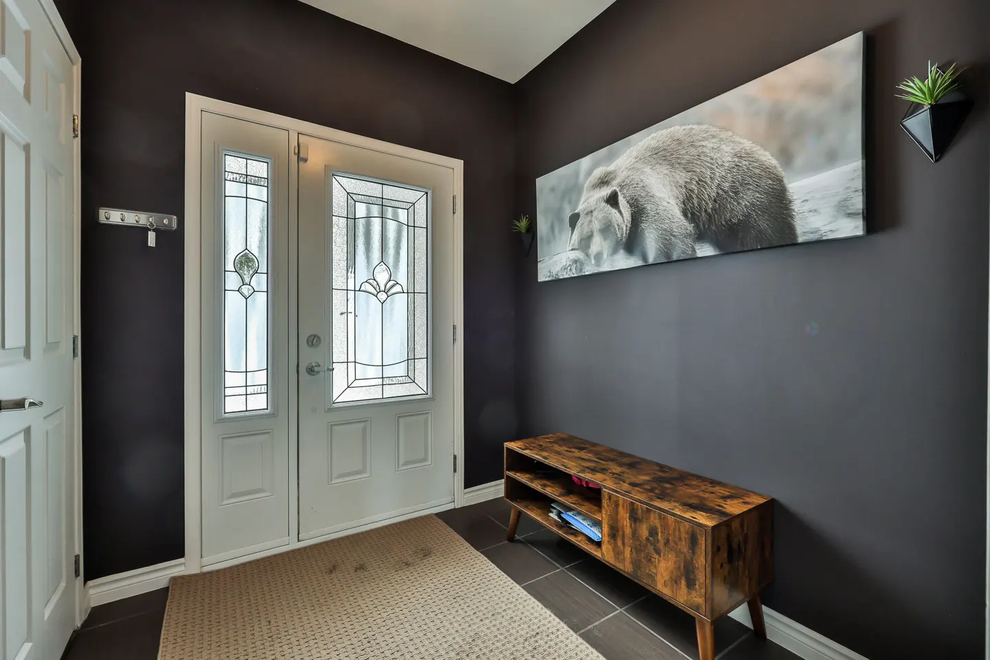 Entryway with white door, bear art, and wood bench. Dark gray walls, tile floor, and a beige rug. A small plant adds a touch of greenery.