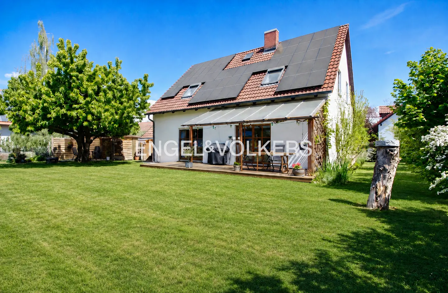A white house with solar panels on the roof, a wooden deck, and a green lawn under a blue sky.