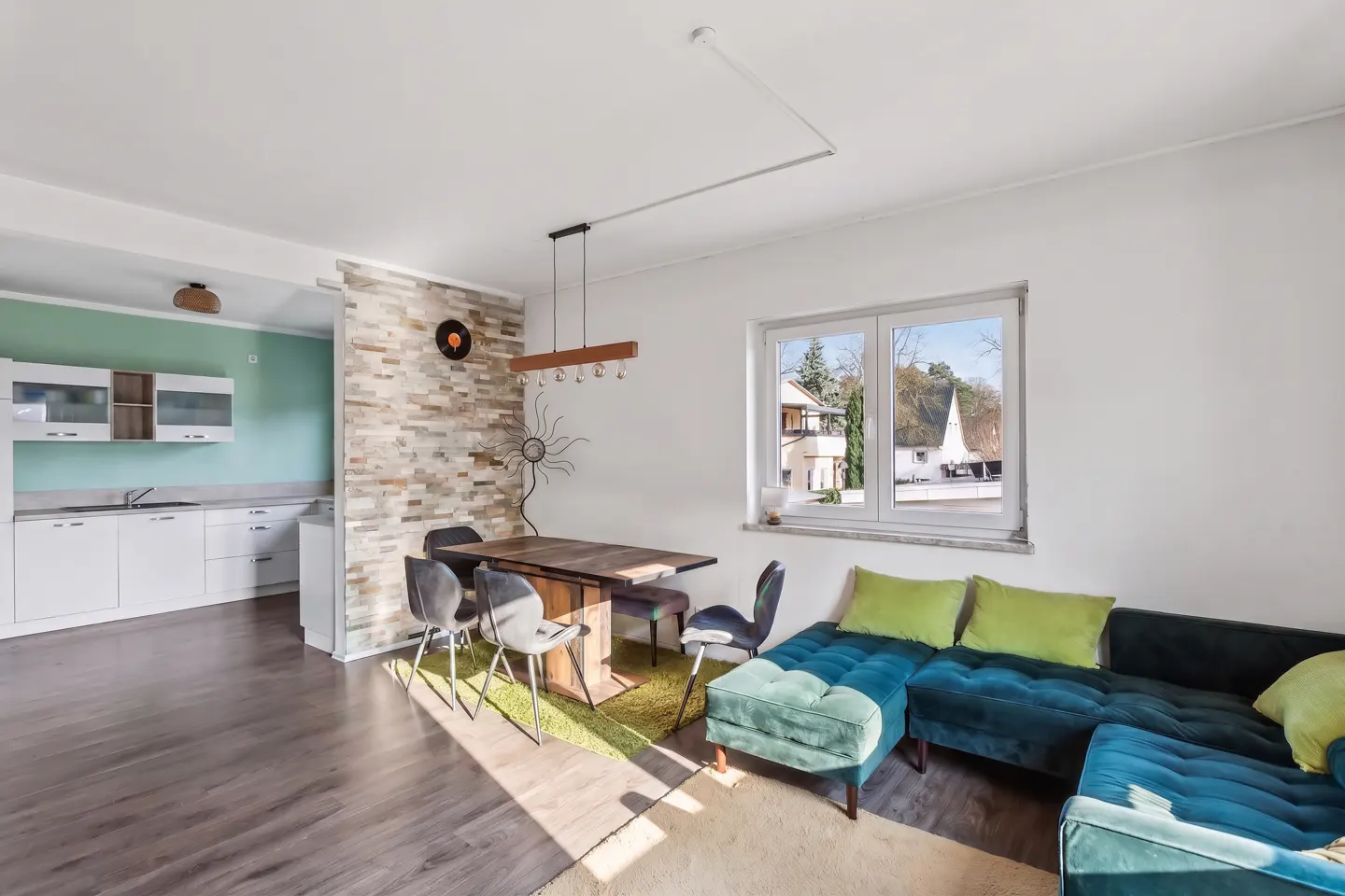 Bright, modern living space with a blue velvet sofa, wooden table, and open kitchen with white cabinets. A window offers a view of the outdoors.