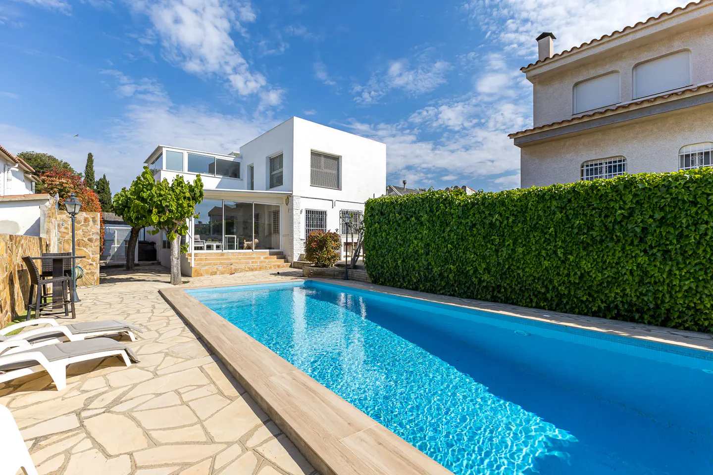 A backyard with a blue swimming pool, stone patio, lounge chairs, and a modern white house under a blue sky.