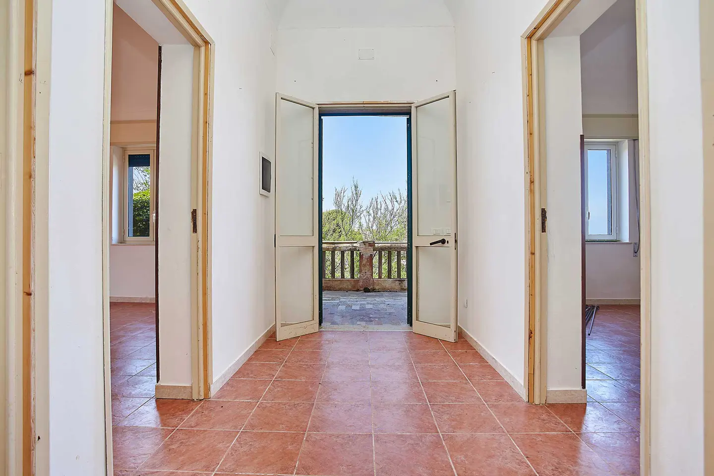 A bright hallway with terracotta tile floors leads to open double doors revealing a sunny balcony with a stone railing.