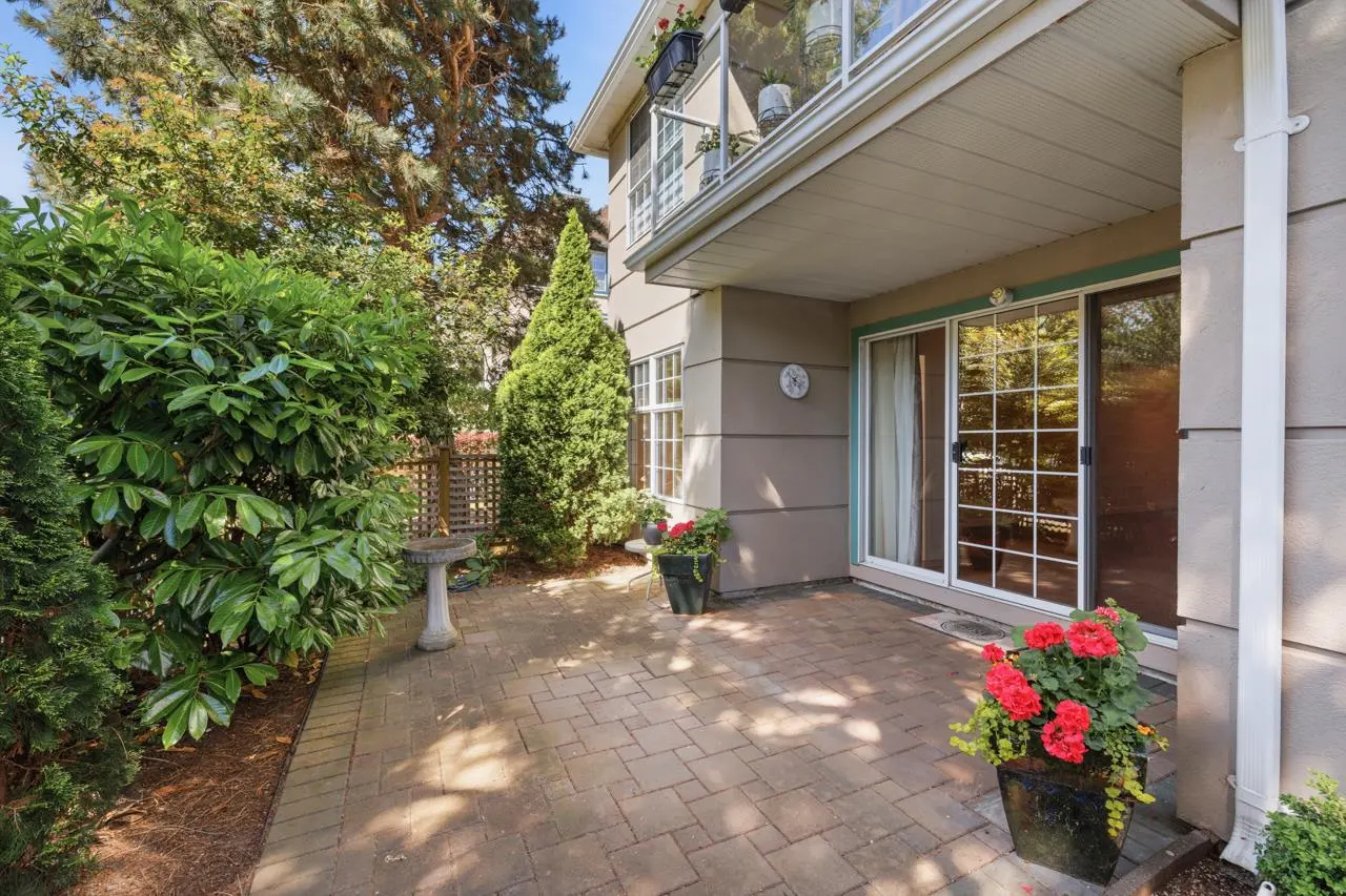 A patio with brick pavers, lush greenery, and a two-story building with sliding glass doors. Red flowers in pots add color.