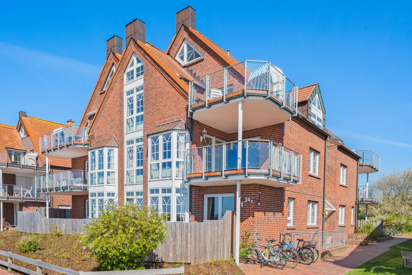 Exterior view of a red brick apartment building with balconies and bikes parked in front. Blue sky background.