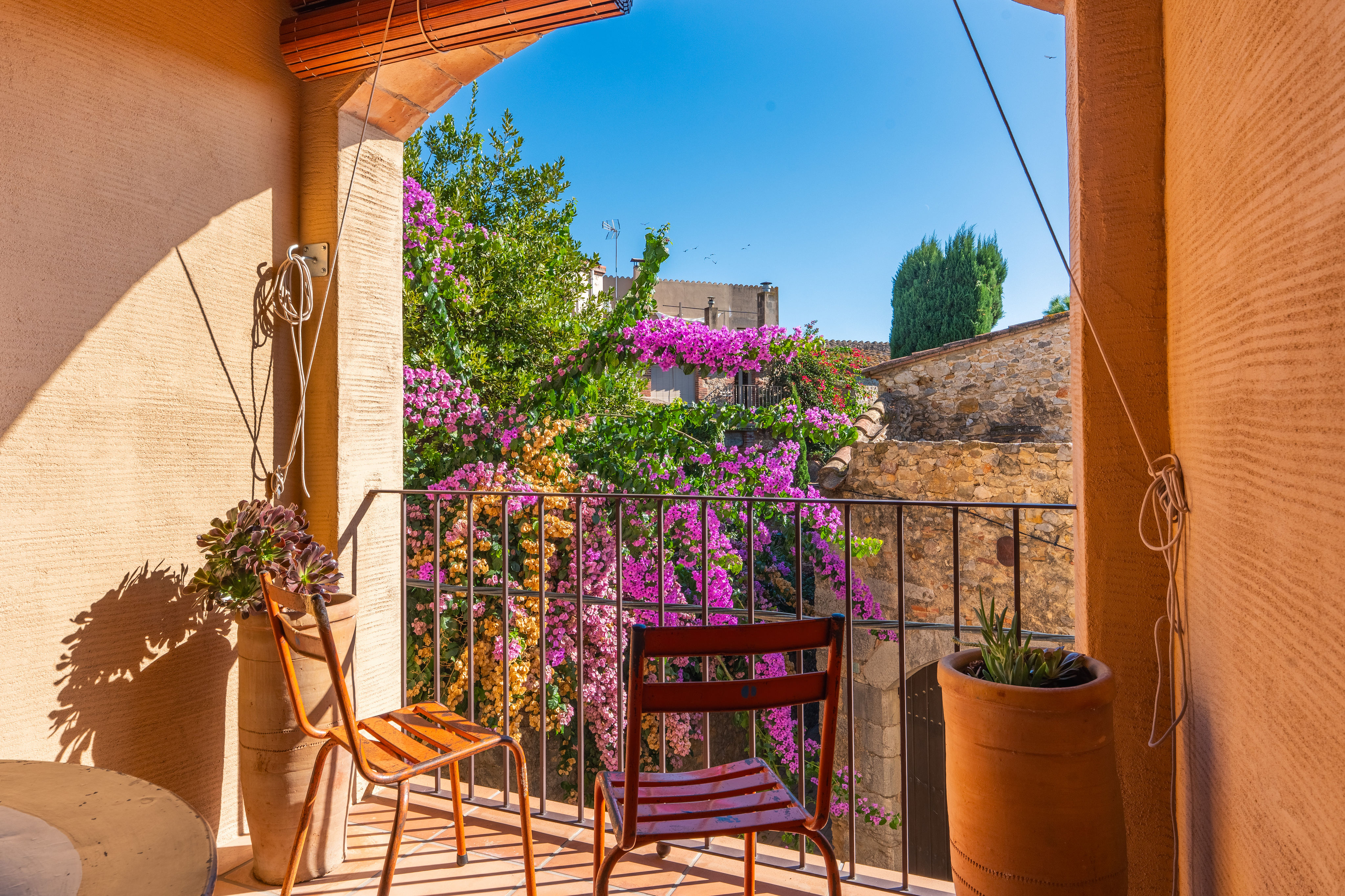 A sunlit balcony with two orange chairs, terracotta pots, and a view of vibrant pink bougainvillea against stone buildings. [AI-generated text]