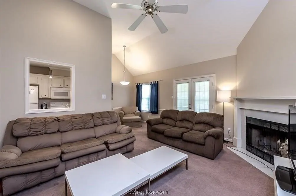 Living room with vaulted ceiling, ceiling fan, brown sofas, white coffee tables, fireplace, and view into kitchen.