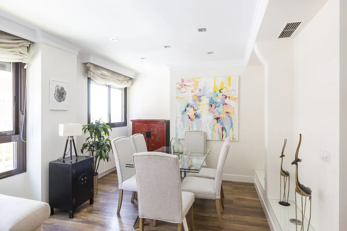 Bright dining room with glass table, upholstered chairs, and colorful abstract art. A red cabinet and black side table add contrast.