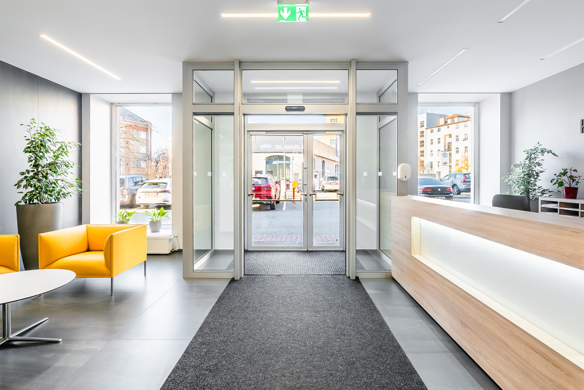 Bright lobby with gray floors, yellow chairs, and a wood reception desk. Glass doors lead outside to a car dealership. [AI-generated text]