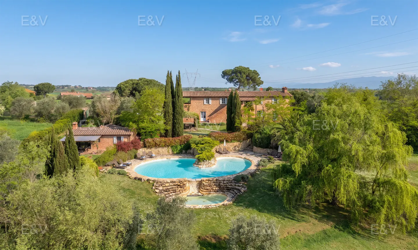 Aerial view of a red brick Tuscan villa with a blue kidney-shaped pool and green trees under a blue sky.