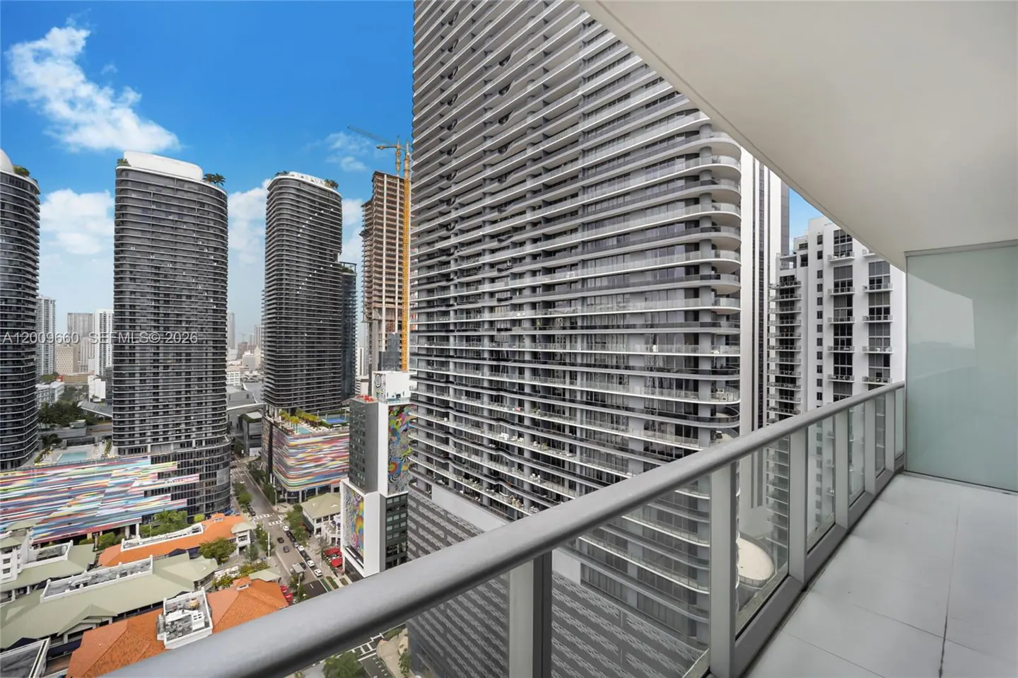 View from a condo balcony overlooking Miami skyscrapers on a sunny day. Gray railings and glass panels line the balcony edge.