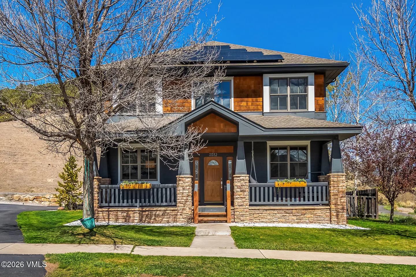 Two-story house with brown shingles, gray siding, stone accents, and solar panels on the roof. A tree stands in the front yard.