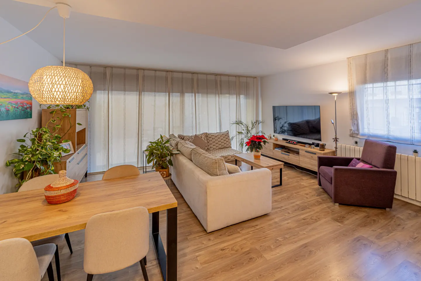 Bright living room with wood floors, dining table, white sofa, TV, and brown armchair. A woven pendant light hangs above the table.