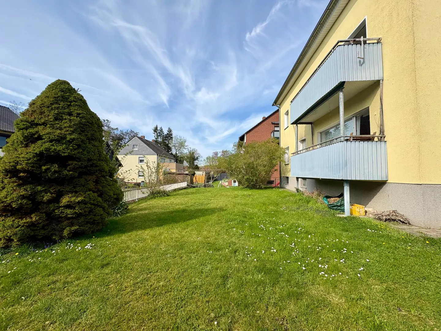 A yellow building with blue balconies overlooks a green lawn with a large evergreen tree and scattered white flowers.