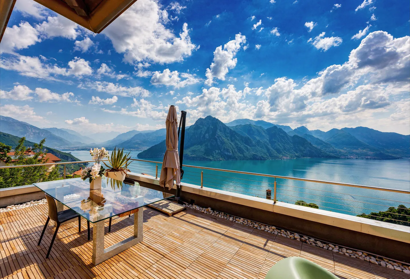 Outdoor patio with glass table, chairs, and umbrella overlooking a lake and mountains under a blue sky with white clouds.
