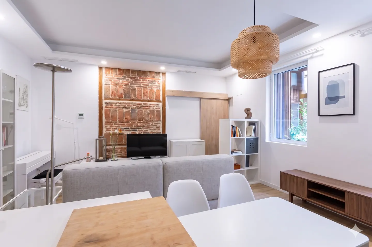 Bright living room with white walls, a brick accent wall, a gray sofa, and a white dining table with chairs. A woven pendant light hangs from the ceiling.