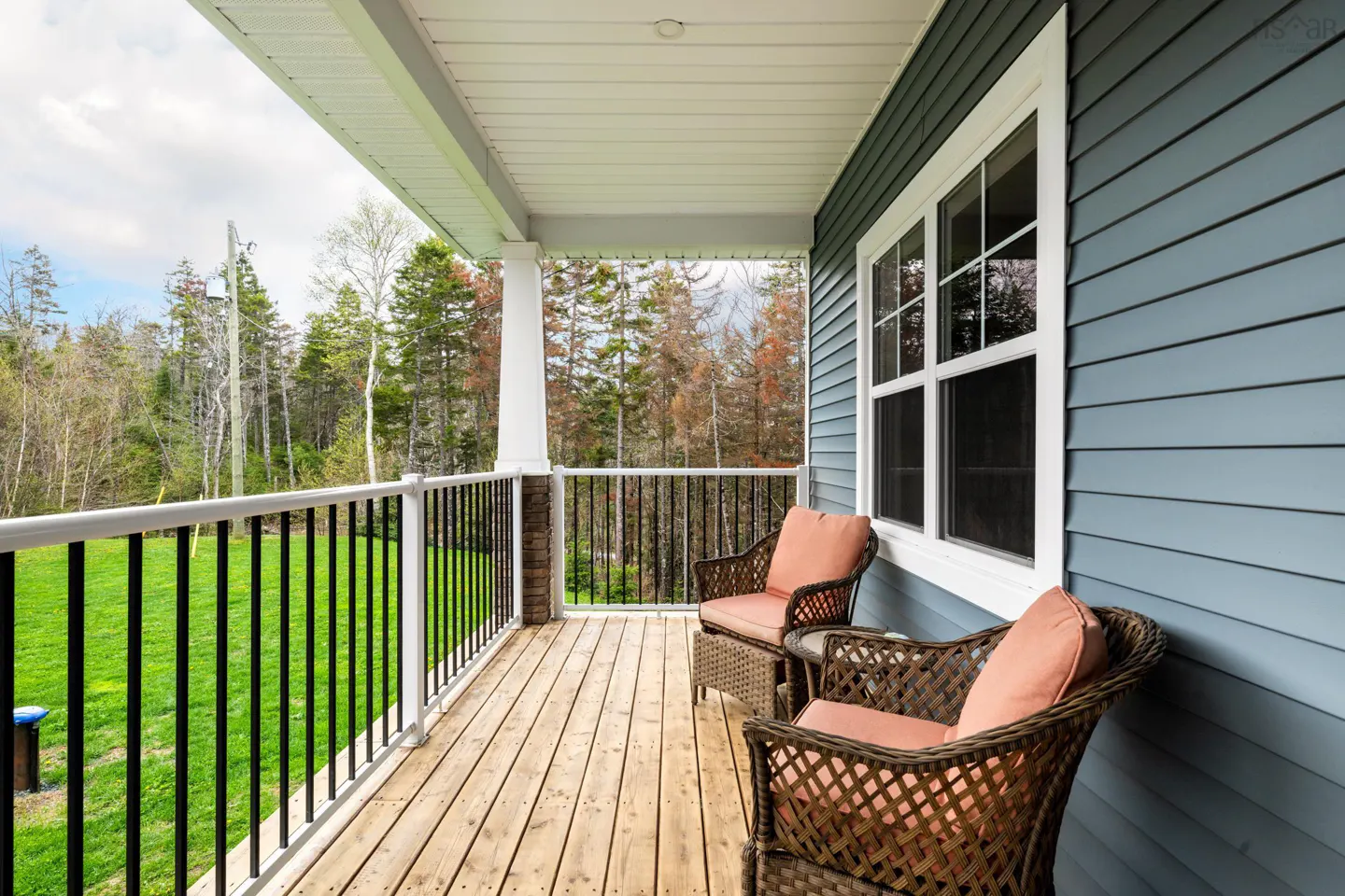 Covered porch with two wicker chairs with coral cushions, a small table, and a view of trees and green grass. The house is blue with white trim.