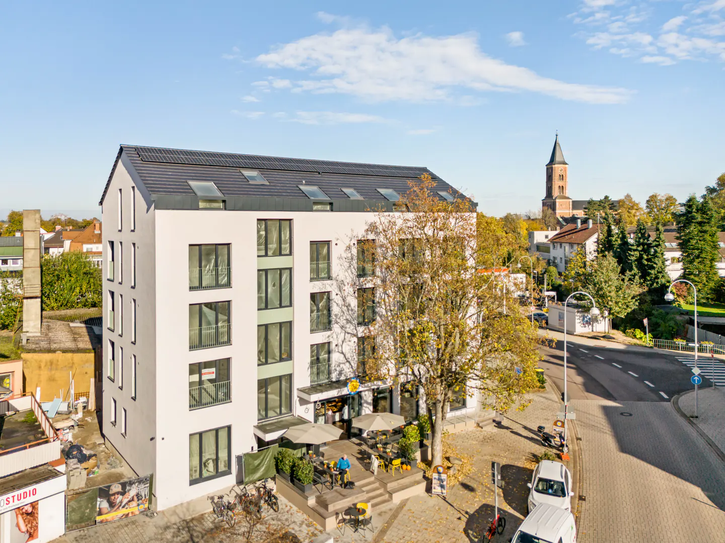 Exterior of a modern white apartment building with a cafe, tables, and a church tower in the background.