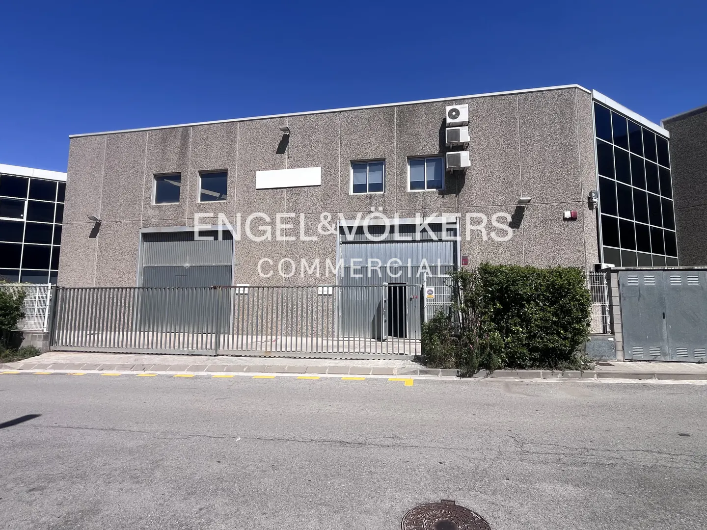 Commercial building with gray concrete walls, metal gates, and an Engel & Völkers Commercial sign under a clear blue sky.