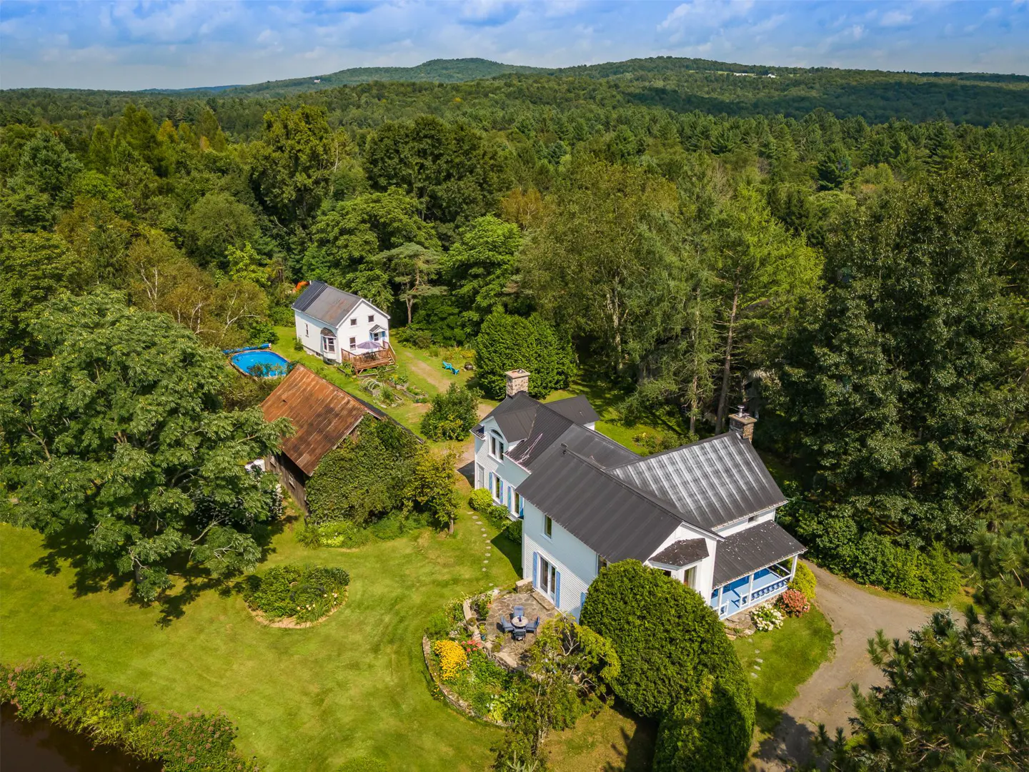 Aerial view of a white farmhouse with a dark roof, surrounded by lush green trees and a mountain backdrop.