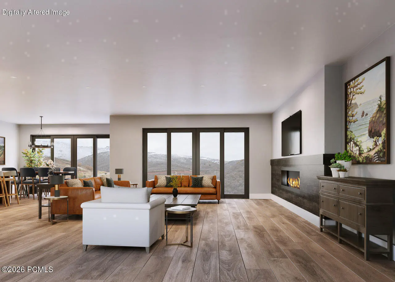 Open-concept living room with hardwood floors, a fireplace, and mountain views through large windows. Brown leather sofas and a white armchair furnish the space.