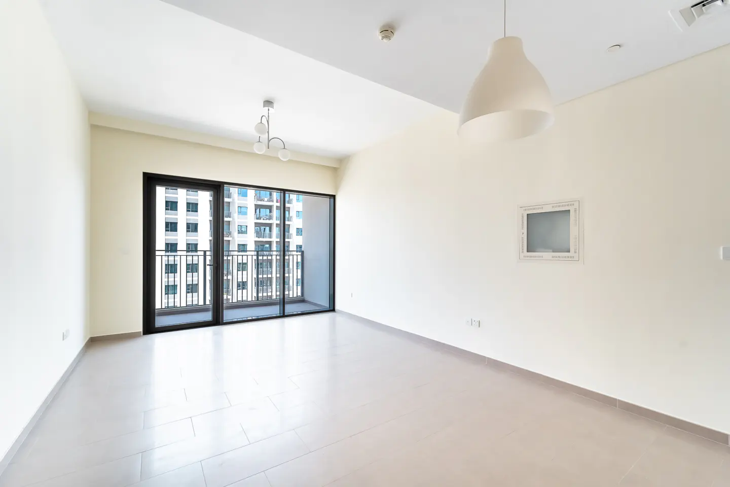 Bright, empty room with beige walls, tile floor, and black-framed sliding glass doors to a balcony with a city view. A white pendant light hangs from the ceiling.