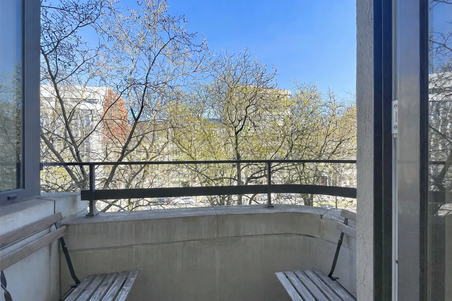 Balcony view with two wooden benches, a black metal railing, and trees against a blue sky.