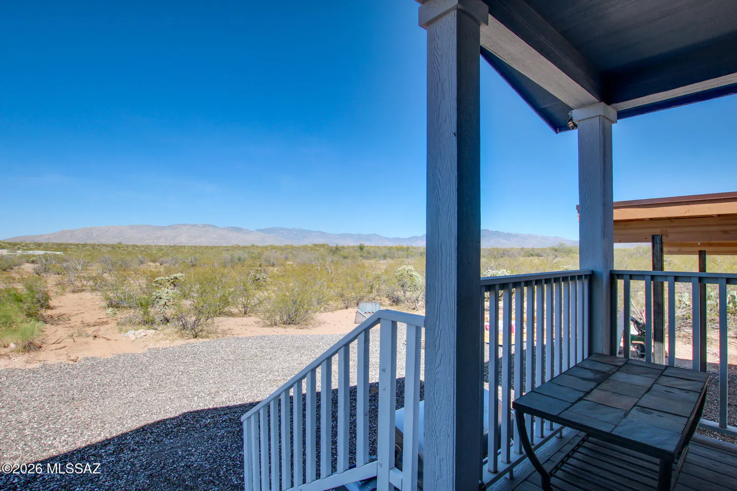 View from a porch with white railings and columns, overlooking desert landscape with mountains under a clear blue sky. A small table sits on the porch.