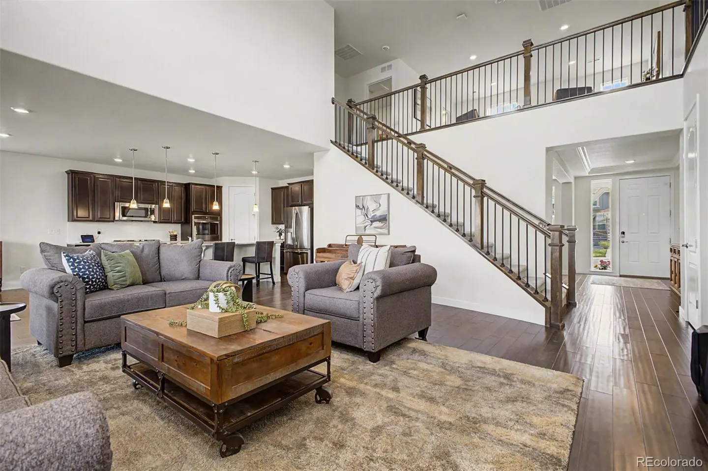 Open-concept living room with gray sofa, wood coffee table, and staircase leading to a second floor. Kitchen in the background.