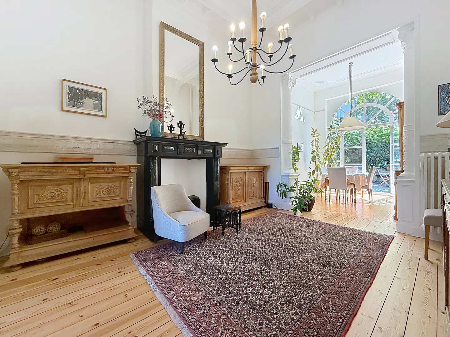 Bright living room with wood floors, a patterned rug, and a black fireplace. A dining area is visible through an arched doorway.