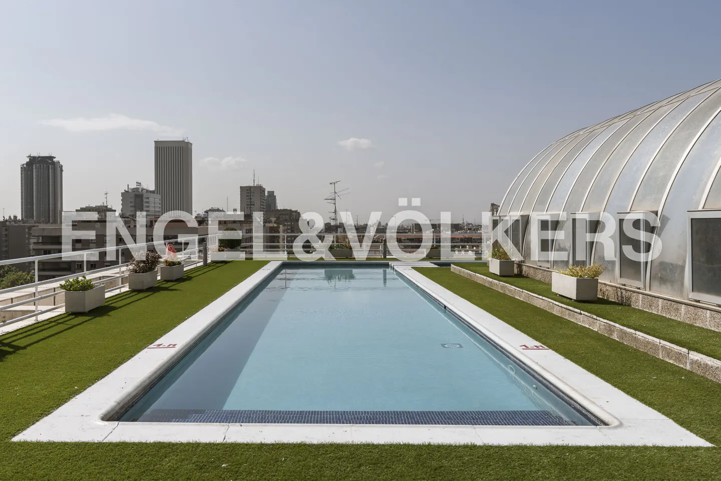 Rooftop swimming pool with blue water surrounded by artificial grass, planters, and a city skyline view under a clear sky.