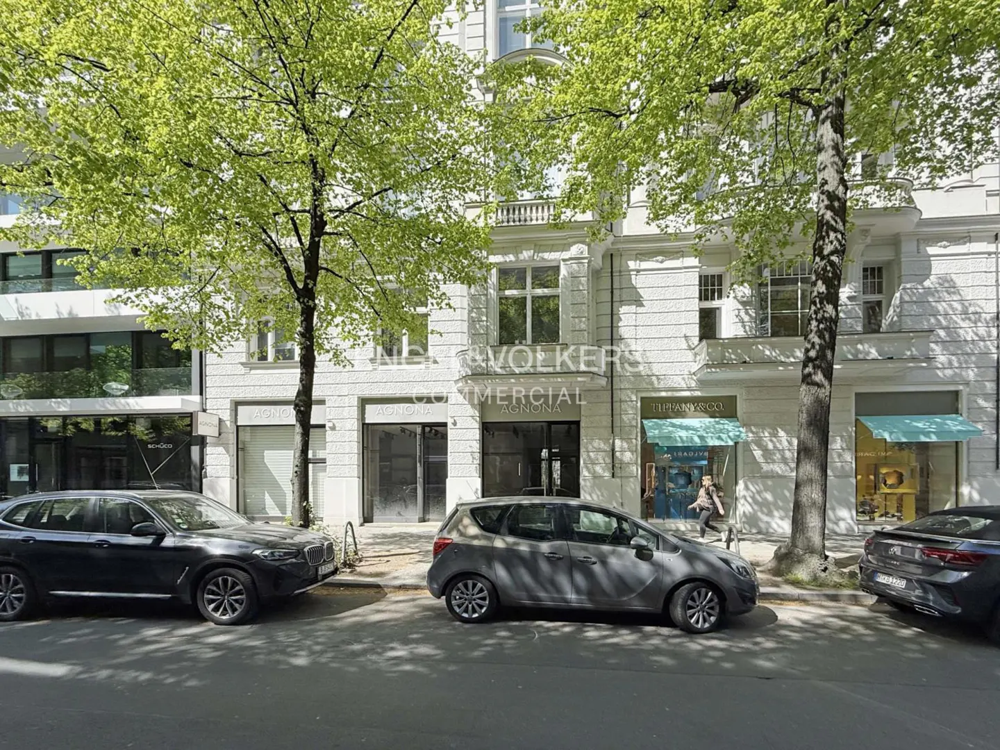 Street view of a white building with "Engel & Volkers Commercial" sign. Cars are parked in front, and trees line the street.