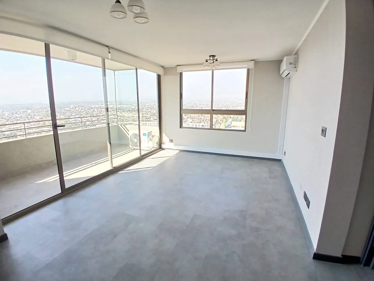 A bright, empty room with gray floors, white walls, and large windows overlooking a city skyline. A balcony is visible through sliding glass doors.
