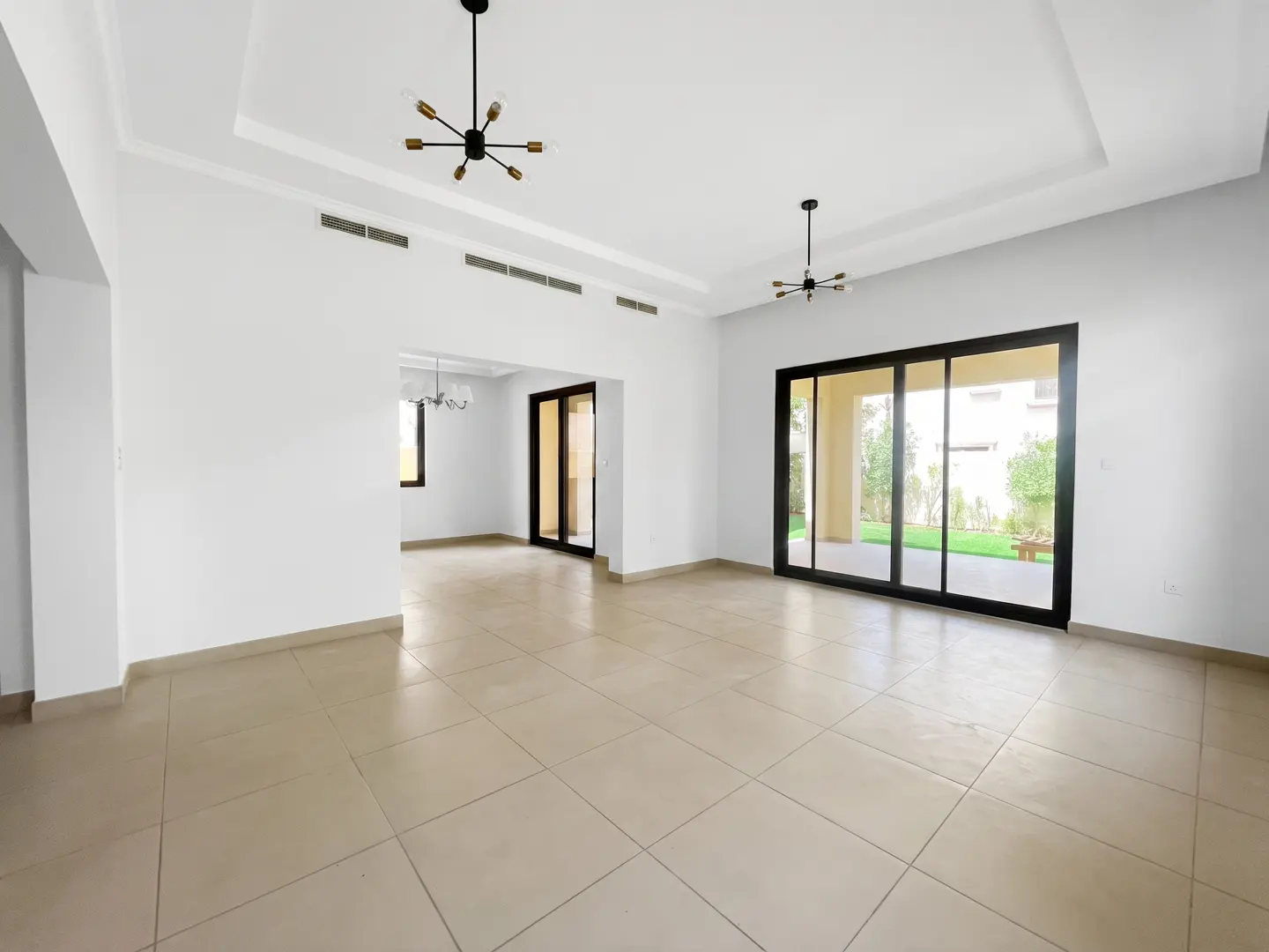 Bright, empty room with white walls, beige tile floor, and black-framed sliding glass doors to a green yard. Two modern chandeliers hang from the ceiling.