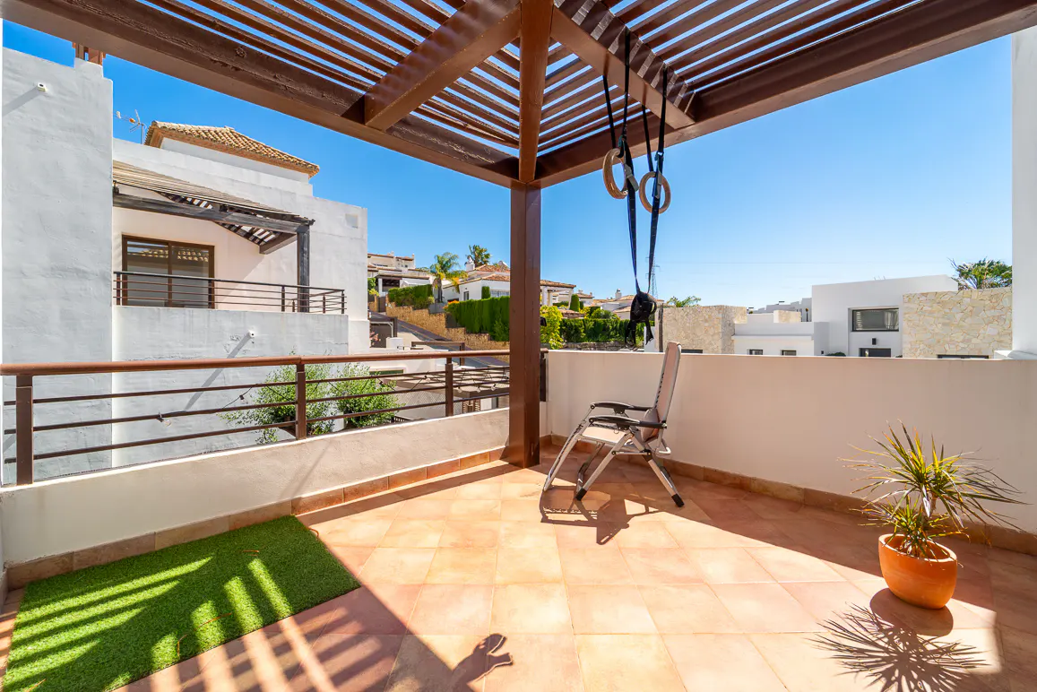 Balcony with terracotta tiles, chair, plant, and gymnastic rings under a wooden pergola. White buildings and blue sky in the background.