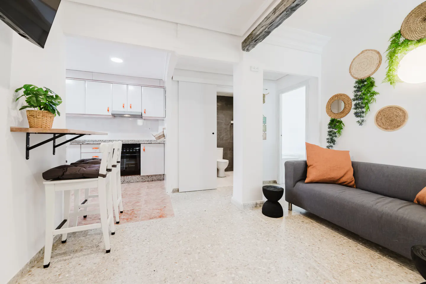 Bright apartment interior with a gray sofa, orange pillow, and straw wall decor. A kitchen with white cabinets and bar stools is visible in the background.