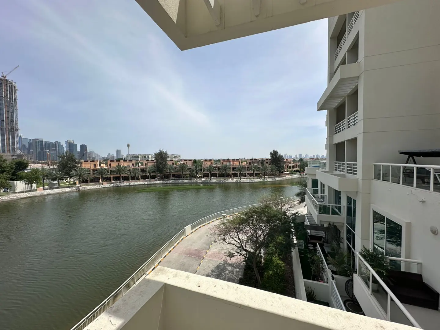 View from a white balcony overlooking a green river, a walkway, and buildings in the distance.
